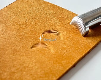 May include: A tan leather sheet featuring two crescent-shaped impressions with fine, radiating lines created by a silver metal stamping tool. The tool is positioned at the edge of the leather, and the text TroyLab is visible in white.