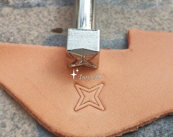 May include: A silver-colored metal leather stamping tool with a square head featuring a four-pointed star design. The tool is positioned above a piece of light tan leather that shows a clear, embossed impression of the same four-pointed star shape.