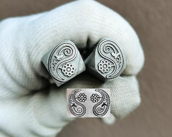 May include: Two silver-colored metal stamps featuring a paisley and flower design, with the text TroyLab visible above. A rectangular impression shows the mirrored paisley and flower pattern created by the stamps, which have square bases.