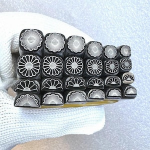 May include: A set of 18 metal stamps featuring a daisy flower design. The stamps are arranged in rows, with the top row featuring a full daisy design and the bottom row featuring a half daisy design.