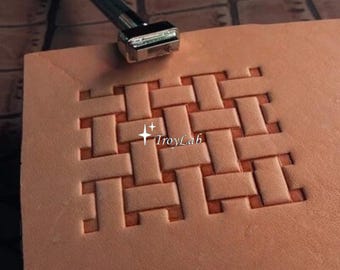 May include: A tan leather square featuring a stamped basketweave pattern and a metal leathercraft tool positioned at the top edge. The center of the stamped design displays the text TroyLab with two small star icons above the word.
