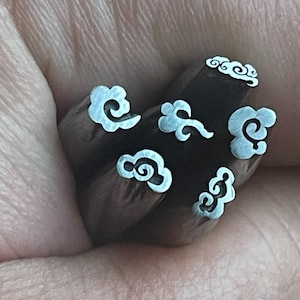 Puede incluir: Un juego de seis sellos metálicos con diseños de nubes. Los sellos se sostienen en una mano y están listos para ser utilizados para manualidades o estampación.