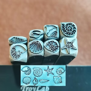 Puede incluir: Un conjunto de sellos metálicos con diseños de conchas marinas, incluyendo un nautilus, una estrella de mar y conchas en espiral. Los sellos están montados sobre bloques de madera. También se ve una placa metálica con el texto "TroyLab" y diseños de conchas.
