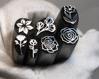 May include: A set of eight metal stamps featuring different floral designs. The stamps are arranged in a circle and are held in a gloved hand. The designs include a rose, a flower, and a leaf.