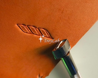 May include: A metal leather stamping tool with a textured handle is pressed into a piece of tan leather, creating a repeating diagonal pattern of three embossed shapes. The text TroyLab is visible near the stamped leather design.
