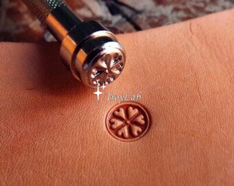 May include: A metal leather stamping tool with a textured handle and a circular tip featuring a six-petaled flower design. The tool is positioned above a piece of tan leather, which shows a clear, stamped impression of the same flower pattern. The text TroyLab is visible.