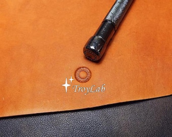 May include: A metal leather stamping tool with a textured grip positioned above a circular impression on a tan leather surface. The text TroyLab is printed in white with two four-pointed star icons next to the circular leather stamp mark.