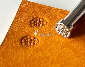 May include: A silver metal leather stamping tool with a circular head featuring a textured pattern of small raised dots. The tool is positioned next to two circular impressions on a piece of tan leather, each showing the same dot pattern. Text reads TroyLab.
