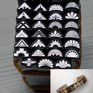 Pode incluir: Um conjunto de 24 carimbos de metal com desenhos geométricos, dispostos em filas. Os carimbos são pretos com desenhos brancos. Uma pulseira de metal dourado com um padrão geométrico é mostrada no canto inferior direito.