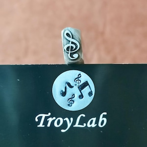 May include: A metal stamp with a treble clef design. The stamp is positioned above a black surface with the text "TroyLab" and a circular graphic of musical notes. The background is a solid brown color.