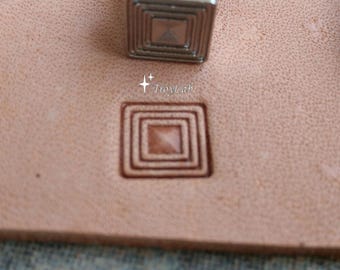 May include: A metal leather stamping tool with a square base featuring concentric square grooves. The tool is positioned above a light tan leather surface that displays a clear, pressed impression of the same square pattern with the text TroyLab.
