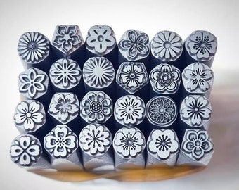Op de afbeelding: Een set van 24 metalen stempels met verschillende bloemmotieven. De stempels zijn gerangschikt in een rasterpatroon en zijn allemaal zilverkleurig.