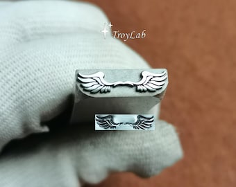 May include: A silver-colored metal stamping tool featuring a raised wing design on its end. The tool is marked with the text TroyLab and includes a small inset showing the stamped impression of the wings, which are symmetrical and detailed.