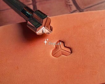May include: A metal leather stamping tool with a three-pronged head positioned above a light brown leather surface. The tool has created a matching three-pronged impression in the leather. The text TroyLab is visible near the impression.