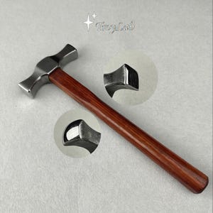 May include: A metal hammer with a polished silver-toned head and a smooth, reddish-brown wooden handle. The head features two distinct striking surfaces, with circular insets showing detailed views of the curved and flat metal ends. TroyLab text.