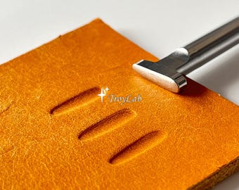 May include: A silver-colored metal leathercraft tool with a flat, rounded tip is positioned on a square piece of textured orange leather. The leather features three parallel, elongated, indented impressions. The text TroyLab is visible.