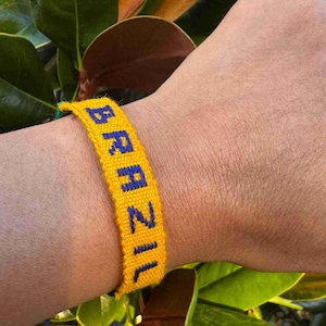 以下が含まれることがあります： 黄色い織りブレスレットで、青色で「BRAZIL」の文字が刺繍されています。 ブレスレットは手首に着用され、背景には緑の葉があります。 ブレスレットは鮮やかなアクセサリーです。