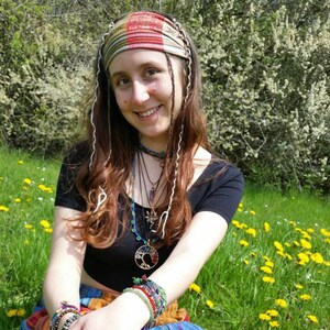 May include: A multi-colored plaid headband with white wavy trim, a black short-sleeved top, and a layered necklace featuring a circular tree of life pendant with multi-colored stones. The ensemble includes several beaded bracelets on the wrists.