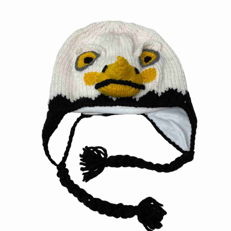 Peut inclure: Chapeau en tricot blanc avec un motif de visage d'aigle noir et blanc. Le chapeau a des rabats d'oreilles noirs et un long cordon tress&eacute; noir avec des glands.