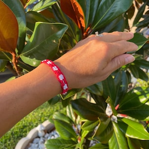 May include: A red and white friendship bracelet with a checkered pattern. The bracelet is made of woven string and is worn on a person's wrist.
