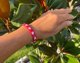May include: A red and white friendship bracelet with a checkered pattern. The bracelet is made of woven string and is worn on a person's wrist.