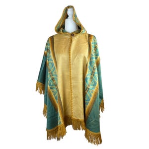 May include: A hooded poncho featuring a central mustard yellow panel flanked by teal and brown geometric patterned side sections. The garment includes a single button closure at the neckline and a mustard yellow fringe trim along the bottom hem.