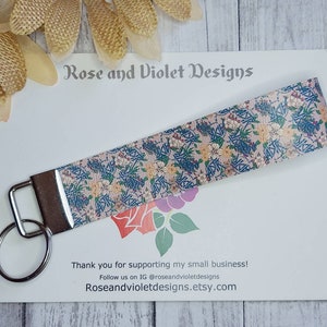 May include: A floral patterned key chain with a silver key ring. The keychain is made of fabric and has a pink, blue, and green floral pattern. The text "Rose and Violet Designs" is printed on a white card behind the keychain.