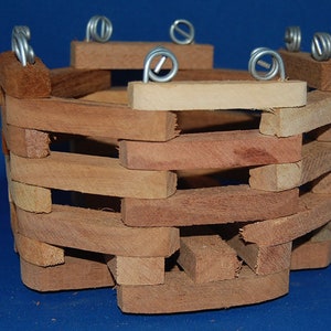 May include: A small, handcrafted wooden basket with a woven design. The basket is made of light and dark brown wood pieces, with silver wire handles attached to the top. The basket is likely used for decorative purposes.