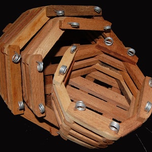 May include: A wooden hanging basket with a geometric design. The basket is constructed from narrow, light brown wooden slats held together by small metal springs. The basket's shape is an irregular octagon, with an open interior.