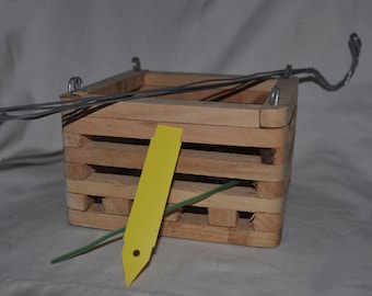May include: A small, square wooden hanging basket with a natural finish. The basket is constructed from horizontal wooden slats and features metal wire hangers. A yellow tag and green wire are also present.