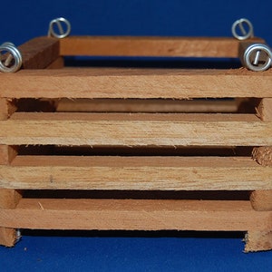 May include: A small, handcrafted wooden crate with a rectangular shape. The crate is constructed from light brown wood and features horizontal slats for the sides. Silver wire loops are attached to the top corners.
