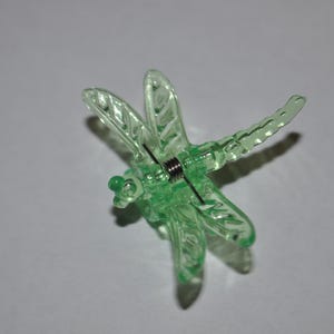 May include: A translucent green dragonfly-shaped clothespin. The wings and body are detailed with vein-like patterns. A silver-colored spring is visible in the center. The clothespin is designed to hold items.