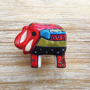 May include: A colourful ceramic elephant figurine with red, white, blue, green and black stripes and polka dots. The elephant is standing on a light wood surface.