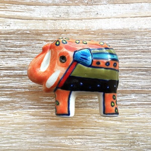 May include: A colourful ceramic elephant figurine with orange, green, blue, black and white paint. The elephant has a whimsical design with dots and stripes.