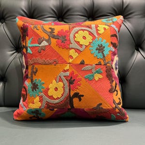 Könnte beinhalten: Quadratisches Zierkissen mit Patchwork-Design. Das Kissen hat eine lebendige Mischung aus orange, rot, gelb und türkis. Das Design umfasst florale und abstrakte Stickmuster. Das Kissen liegt auf einer dunkelgrauen, getufteten Oberfläche.