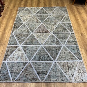 Puede incluir: Una alfombra de patchwork con un patrón geométrico de triángulos en tonos azules, grises y marrones. La alfombra tiene un borde blanco y está hecha de materiales reciclados.