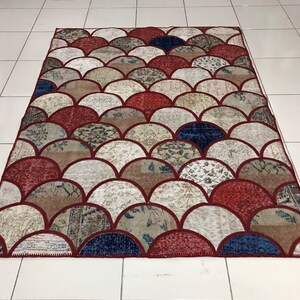 May include: A colourful patchwork rug with a scale pattern in shades of red, brown, and white. The rug is made from recycled materials and has a unique, handmade look.