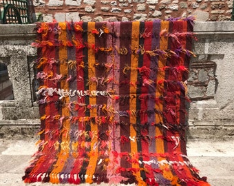 May include: A colorful striped rug with a fringe border. The rug is made of wool and has a variety of colors, including red, orange, yellow, brown, and purple.