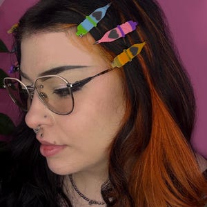 May include: A person with black hair, orange highlights, and three colorful hair clips. The clips are shaped like pencils with heart details, in shades of green, blue, pink, purple, and yellow. The person is wearing glasses with a silver frame.