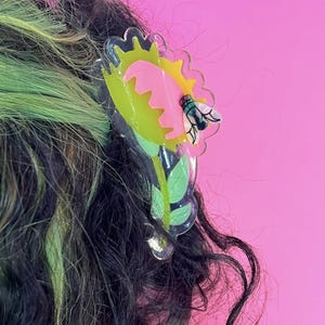 May include: A clear, plastic hair clip shaped like a Venus flytrap. The clip features a yellow and pink flower with a green fly and leaves. The clip is set against a pink background and is placed in dark and green hair.