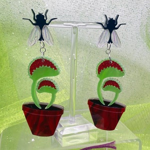 May include: A pair of earrings featuring a green Venus flytrap with red accents and a black fly on top. The flytrap is in a red pot.