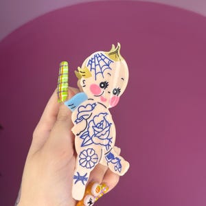 Könnte beinhalten: Cremefarbene, cherubförmige Brosche mit blauen Tattoo-Designs, darunter eine Rose und ein Spinnennetz. Der Cherub hat rosa Wangen, eine blaue Schleife und kleine Flügel. Die Brosche wird in einer Hand mit farbenfroher Nagelkunst gehalten.