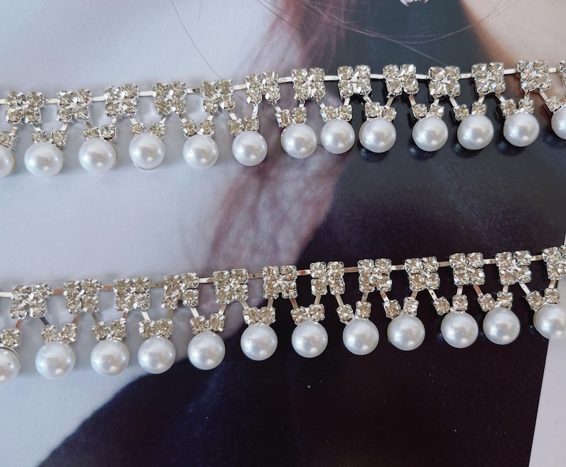 Peut inclure: Deux rang&eacute;es parall&egrave;les de garniture d&eacute;corative compos&eacute;es d'une cha&icirc;ne en m&eacute;tal argent&eacute; sertie de strass transparents et brillants. Chaque segment de strass est reli&eacute; &agrave; une petite perle ronde blanche qui pend du bord inf&eacute;rieur.