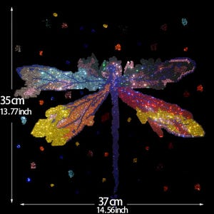 以下が含まれることがあります： 黒い背景に、幅35cm、長さ37cmのカラフルな宝石付きトンボのデザイン。羽は黄色、オレンジ、赤、青、緑などのグラデーションカラーで表現されています。デザインはキラキラとしたディテールで装飾されています。