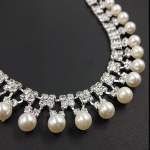 Peut inclure: Un collier argent&eacute; compos&eacute; d'une s&eacute;rie de strass carr&eacute;s transparents dispos&eacute;s en grille, avec des perles blanches rondes suspendues au bord inf&eacute;rieur de chaque groupe de strass, cr&eacute;ant un effet superpos&eacute; et pendant.