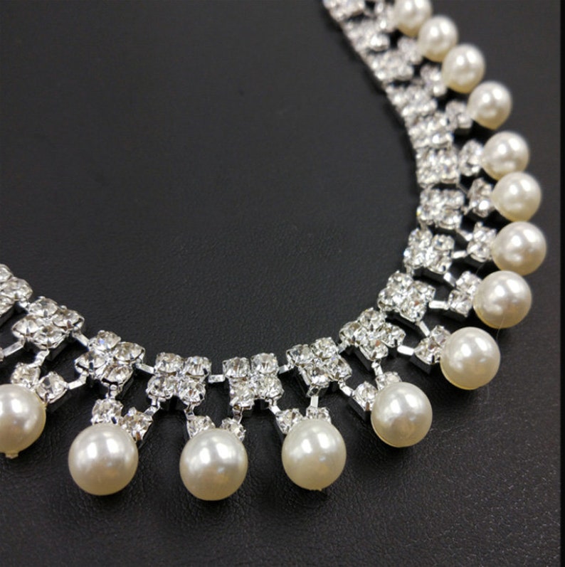 Peut inclure: Un collier argent&eacute; compos&eacute; d'une s&eacute;rie de strass carr&eacute;s transparents dispos&eacute;s en grille, avec des perles blanches rondes suspendues au bord inf&eacute;rieur de chaque groupe de strass, cr&eacute;ant un effet superpos&eacute; et pendant.