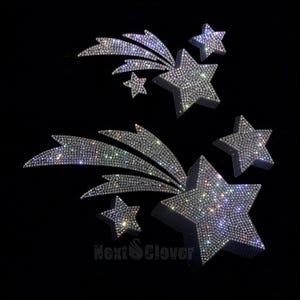 May include: Two shooting star motifs covered in clear, reflective rhinestones that create a sparkling effect. Each design features a large five-pointed star with a trailing tail and smaller accent stars. The text NextClover is visible at the bottom.
