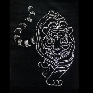 Può includere: Una superficie in tessuto nero con un disegno di tigre creato con piccoli strass bianchi riflettenti. La tigre è raffigurata in una posa frontale con la coda curva verso l'alto, mettendo in risalto il dettagliato motivo a strisce.