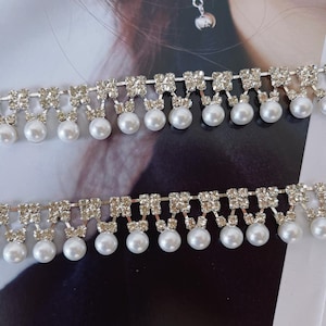 Peut inclure: Deux bandes de garniture d&eacute;corative compos&eacute;es d'une rang&eacute;e horizontale de strass carr&eacute;s transparents reli&eacute;s par des maillons m&eacute;talliques argent&eacute;s, avec une rang&eacute;e de perles blanches rondes suspendues &agrave; chaque carr&eacute; de strass.