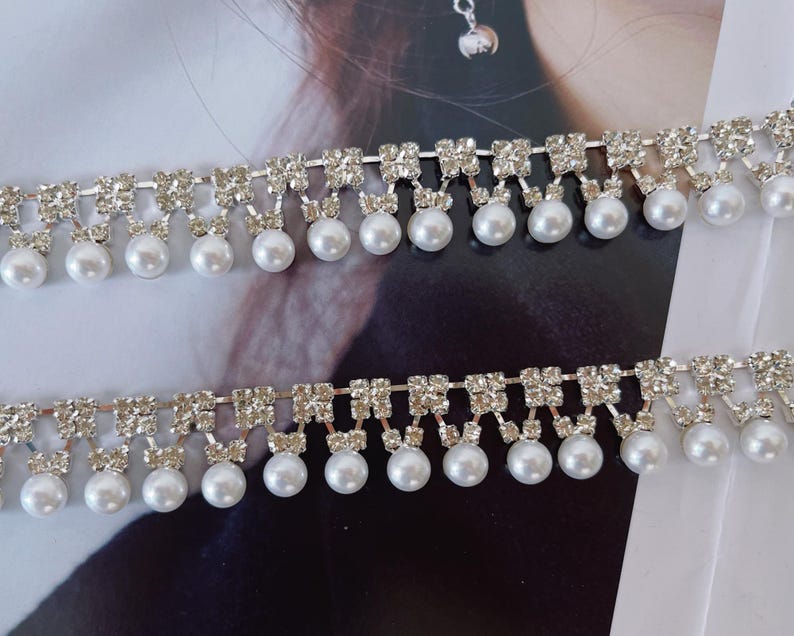 Peut inclure: Deux bandes de garniture d&eacute;corative compos&eacute;es d'une rang&eacute;e horizontale de strass carr&eacute;s transparents reli&eacute;s par des maillons m&eacute;talliques argent&eacute;s, avec une rang&eacute;e de perles blanches rondes suspendues &agrave; chaque carr&eacute; de strass.